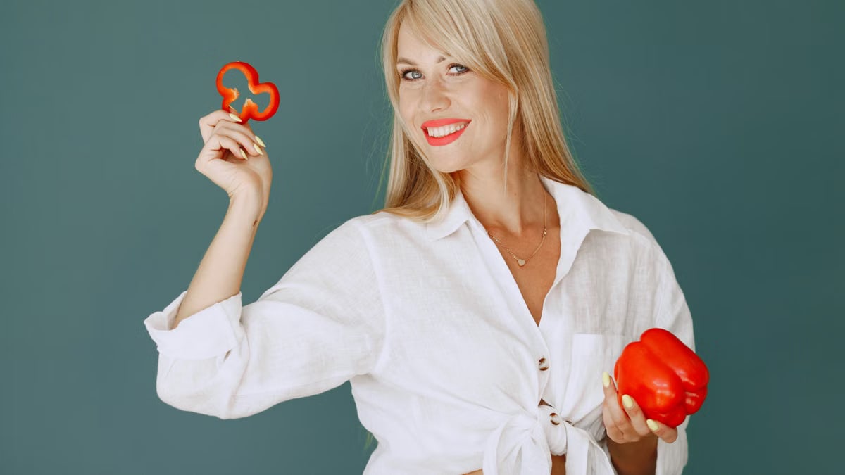 Blonde woman in white shirt smiling while holding a fresh red bell pepper slice.